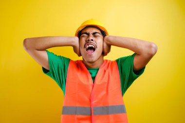 Güvenlik kaskı takan Asyalı inşaat işçisi ve yüksek sesli müzik sesi için kulaklarını parmaklarıyla kapatan üniformalı bir işçi. Sağır kavramı.