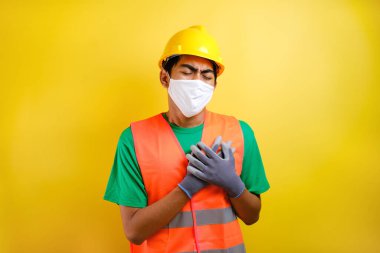 Güvenlik kaskı ve koruyucu maske takan Asyalı inşaat işçisi göğsünde ağrı hissederken sarı arka plana bastırıyor.