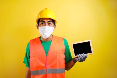 Koronavirüse karşı koruyucu maske takan Asyalı işçinin portresi, sarı arka planda küçük bir karatahta tutuyor.