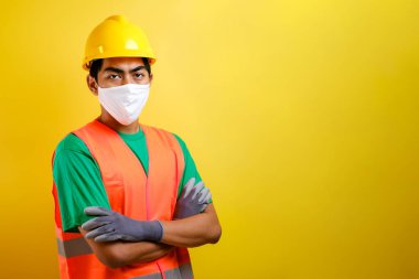 Maskeli ve güvenlik yeleği giyen Asyalı müteahhit sarı arka plana karşı güven jesti yaparak kolunu çarpmış.