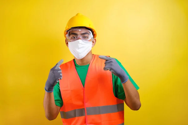 Asian construction worker pointing at his protective mask and savety helmet invites to prioritize health and safety against yellow background
