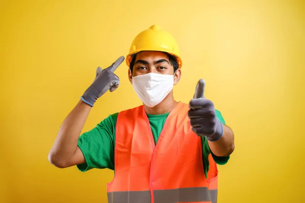 Asian construction worker pointing at his protective mask and savety helmet invites to prioritize health and safety against yellow background