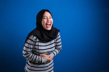 Asian muslim woman laughing hard while holding her belly, happy excited expression against blue background