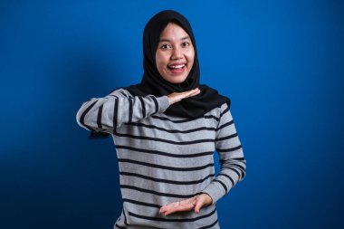 Portrait of Asian young happy muslim woman presenting something between her hands, against blue background with copy space