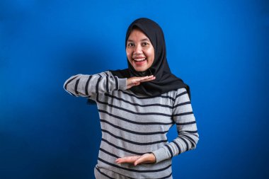 Portrait of Asian young happy muslim woman presenting something between her hands, against blue background with copy space