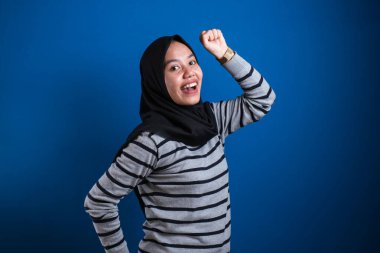 Portrait of a funny young Asian muslim girl wearing hijab dancing happily joyful expressing celebrating good news victory winning success gesture, smiling positive excited emotion isolated on blue background