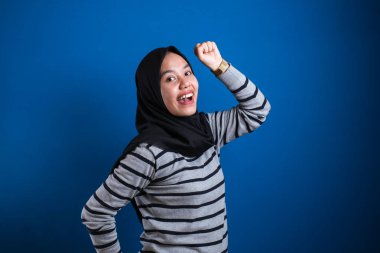 Portrait of a funny young Asian muslim girl wearing hijab dancing happily joyful expressing celebrating good news victory winning success gesture, smiling positive excited emotion isolated on blue background