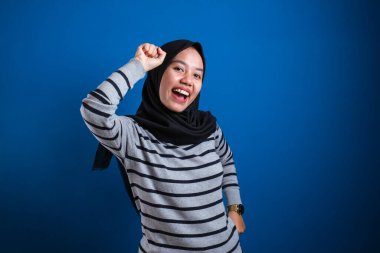 Portrait of a funny young Asian muslim girl wearing hijab dancing happily joyful expressing celebrating good news victory winning success gesture, smiling positive excited emotion isolated on blue background