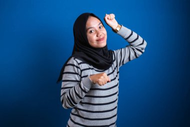 Portrait of a funny young Asian muslim girl wearing hijab dancing happily joyful expressing celebrating good news victory winning success gesture, smiling positive excited emotion isolated on blue background