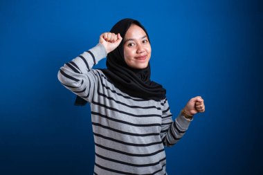 Portrait of a funny young Asian muslim girl wearing hijab dancing happily joyful expressing celebrating good news victory winning success gesture, smiling positive excited emotion isolated on blue background