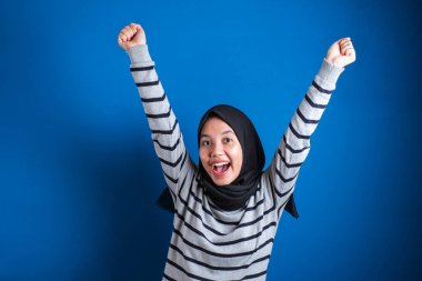 Büyük kazanç. Başörtülü neşeli Asyalı öğrenci kız ellerini kaldırarak başarıyı kutluyor ve mavi arka planda açık alanda yumruklarını sıkıyor.