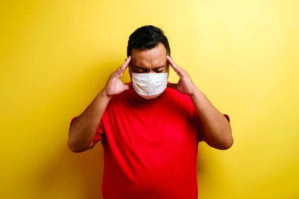Portrait of young ill man in medical protective mask on face suffering from headache and weakness, isolated on yellow background