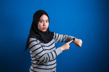 Portrait of muslim student college girllooked angry while showing her wrist watch, late time concept, isolated on blue background
