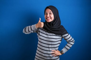 Portrait of happy smiling Asian muslim woman shows OK finger sign gesture, against blue background
