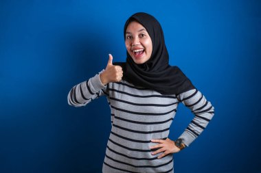 Portrait of happy smiling Asian muslim woman shows OK finger sign gesture, against blue background