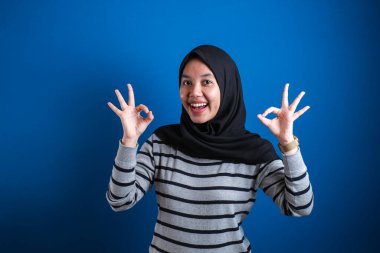 Portrait of happy smiling Asian muslim woman shows OK finger sign gesture, against blue background