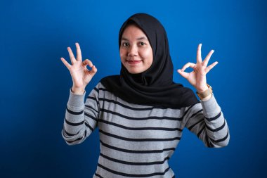 Portrait of happy smiling Asian muslim woman shows OK finger sign gesture, against blue background