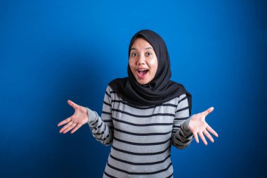 Asian muslim girl wearing hijab shows surprised or shocked expression with open mouth, close up facial expression over blue background