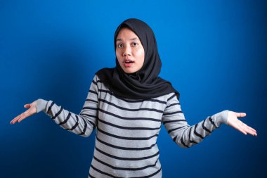 Young beautiful asian muslim student  shrugs her arms, makes gesture of I don't know, can't help anything against blue backgound