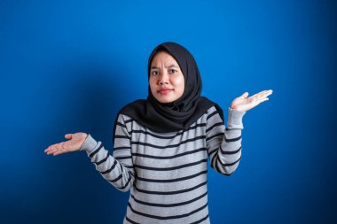 Young beautiful asian muslim student  shrugs her arms, makes gesture of I don't know, can't help anything against blue backgound