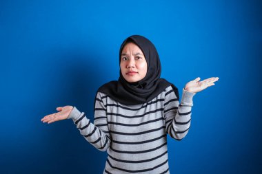 Young beautiful asian muslim student  shrugs her arms, makes gesture of I don't know, can't help anything against blue backgound