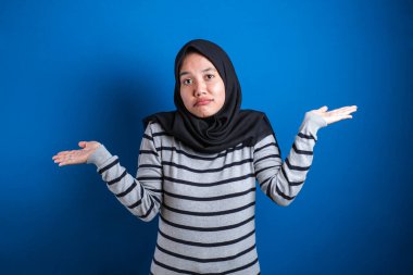 Young beautiful asian muslim student  shrugs her arms, makes gesture of I don't know, can't help anything against blue backgound