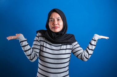 Young beautiful asian muslim student  shrugs her arms, makes gesture of I don't know, can't help anything against blue backgound