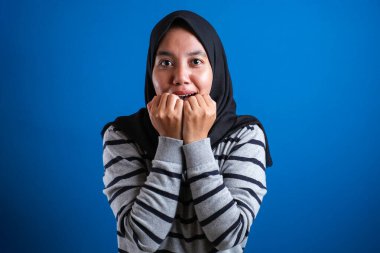 Asyalı Müslüman kadın, mavi arka planda tırnaklarını yerken korkmuş görünüyor.