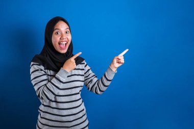 Portrait of Asian young happy asian muslim student girl smiling and pointing to presenting something on her side, with copy space
