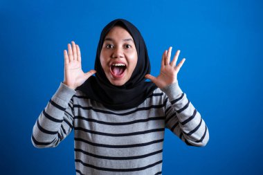 Portrait of cute young Asian muslim college student girl wearing hijab shows surprised or shocked expression with open mouth, close up happy facial expression against blue background