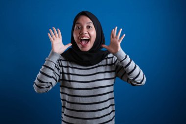 Portrait of cute young Asian muslim college student girl wearing hijab shows surprised or shocked expression with open mouth, close up happy facial expression against blue background