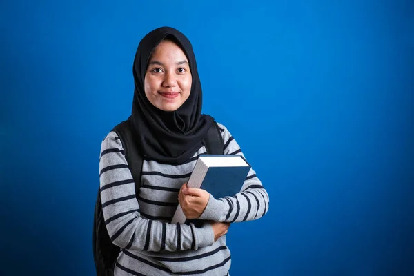 Asian muslim college student girl wearing hijab smiling friendly holding big book against blue background