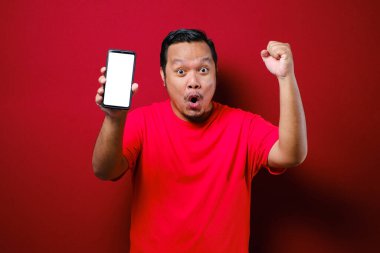 Çekici genç Asyalı adamın telefonunda iyi haberler var. Kırmızı arka plana karşı gülen mutlu bir gülücük.