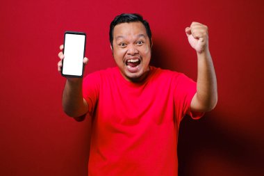 Çekici genç Asyalı adamın telefonunda iyi haberler var. Kırmızı arka plana karşı gülen mutlu bir gülücük.