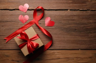 Kırmızı kurdeleli hediye kutusunun seçici odak noktası ve kırsal ahşap masa üzerinde aşk şekilli kağıt, üst görünüm düz.