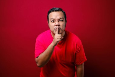 Dudaklarının yanında parmak tutan, sessiz kalmak ve mahremiyetini korumak isteyen ciddi ve katı bir adam. Kırmızı arkaplanda kapalı stüdyo çekimleri