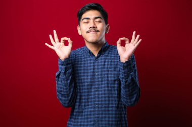 İzole edilmiş kırmızı arka fonun üzerinde dikilen rahat ve gülümseyen genç Hintli adam parmaklarıyla meditasyon hareketi yapıyor. Yoga konsepti.