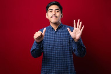 Yakışıklı, gündelik giysiler giymiş altı numarayı gösterip işaret eden genç adam kırmızı arka planda mutlu ve kendinden emin bir şekilde gülümsüyor.
