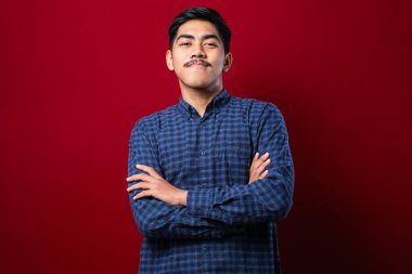 Kırmızı arka plan üzerine gündelik tişört giyen genç yakışıklı kollarını kavuşturup kameraya bakarak gülümsüyor. pozitif kişi.