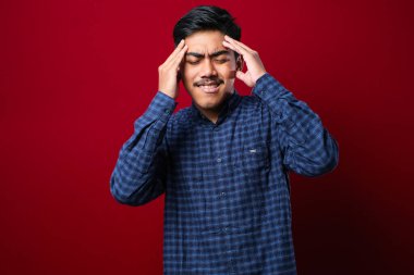 Genç yakışıklı Asyalı bir adam, günlük gömleğini giyip izole edilmiş kırmızı arka planda dikiliyor. Başı ağrıyor çünkü stres altında. Migren ağrısı..