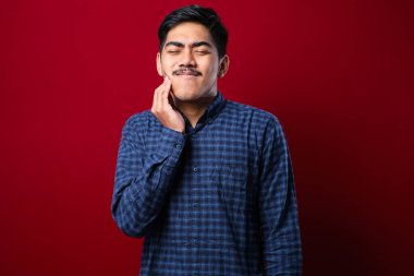 İzole edilmiş kırmızı arka plan üzerinde dikilen genç yakışıklı bir adam diş ağrısı ya da diş ağrısı yüzünden ağız ve acı dolu bir ifade ile dokunuyor. Dişçi