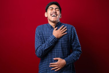 Genç yakışıklı Asyalı çocuk, günlük gömleğini giymiş, kırmızı arka planda dikiliyor, gülümsüyor ve kahkahalarla gülüyor..