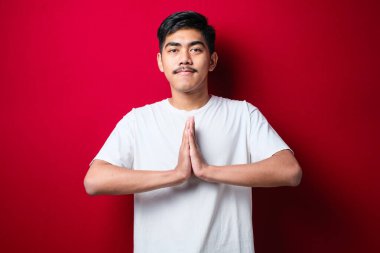 Kırmızı arka planda selamlama ve hoş geldin jestleri içeren günlük beyaz tişört giyen genç Asyalı adam.