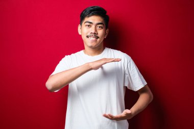 Kırmızı arka planda beyaz tişört giyen yakışıklı Asyalı adam. Ellerinde büyük ve büyük boy işareti, ölçü sembolü var. Kameraya bakıp gülümsüyor. ölçüm kavramı.