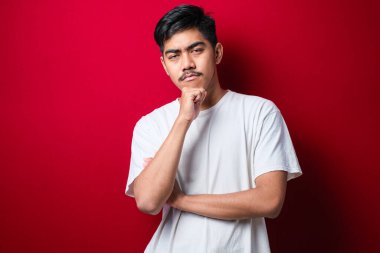 Kırmızı arka planda beyaz tişört giyen yakışıklı Asyalı adam kameraya gülümseyen, kollarını kavuşturup çenesine kaldırmış. Olumlu düşünüyorum..