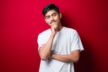 Kırmızı arka planda beyaz tişört giyen yakışıklı Asyalı adam kameraya gülümseyen, kollarını kavuşturup çenesine kaldırmış. Olumlu düşünüyorum..