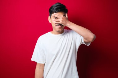 Soyutlanmış kırmızı arka plan üzerinde genç yakışıklı bir adam. Şoka girmiş, yüzünü ve gözlerini kapamış, utangaç ifadesiyle parmaklarından bakıyor..