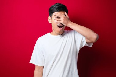 Soyutlanmış kırmızı arka plan üzerinde genç yakışıklı bir adam. Şoka girmiş, yüzünü ve gözlerini kapamış, utangaç ifadesiyle parmaklarından bakıyor..