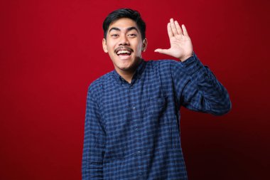 Kırmızı arka plan üzerinde duran beyaz tişörtlü genç yakışıklı Asyalı. Mutlu ve güler yüzlü, dostça bir jest yapıyor.