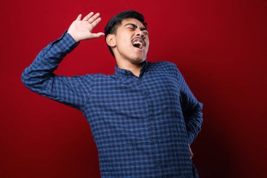 Beyaz tişörtlü genç yakışıklı Asyalı. Kırmızı arka planda duruyor. Yorgun, rahat, uykulu ve sabahın erken saatlerinde esniyor.